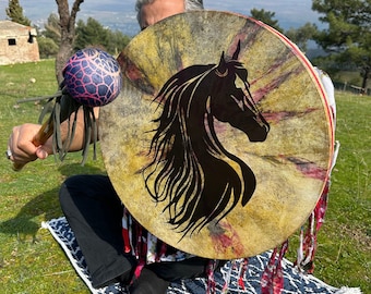 May include: A circular drum featuring a black horse head silhouette. The drum has a natural, aged look with red and yellow detailing. A mallet with a purple and blue patterned head is held in the left hand. The drum is decorated with red and white fabric.