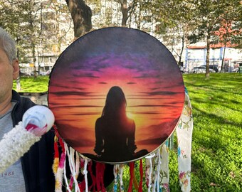 Puede incluir: Un tambor redondo con un diseño de puesta de sol, que representa una silueta en posición de meditación. El diseño del tambor utiliza tonos de púrpura, naranja y amarillo. Telas y borlas coloridas adornan el borde del tambor.