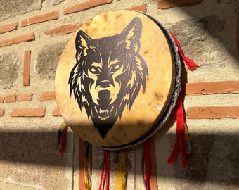 Puede incluir: Un tambor redondo con un diseño de lobo. El parche del tambor es de color beige con una silueta de lobo negro. El tambor tiene un marco de madera con cintas de tela rojas, amarillas y naranjas colgando de la parte inferior. El tambor está colgado en una pared de ladrillo.