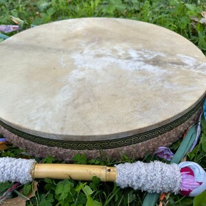 May include: A round, natural-toned drum with a light brown hide stretched over a circular frame. The frame is wrapped in a textured, light brown material with a decorative green and gold border. A drumstick with a wooden handle and gray, fluffy ends rests beside it.