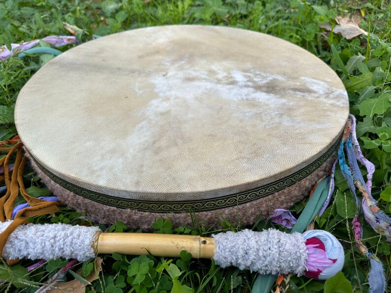 May include: A round, natural-toned drum with a light brown hide stretched over a circular frame. The frame is wrapped in a textured, light brown material with a decorative green and gold border. A drumstick with a wooden handle and gray, fluffy ends rests beside it.