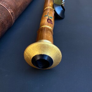 May include: A handcrafted wooden wind instrument with a gold-colored mouthpiece and a rose design. The instrument is accompanied by a brown leather case and a small bottle. The instrument's body is made of bamboo.