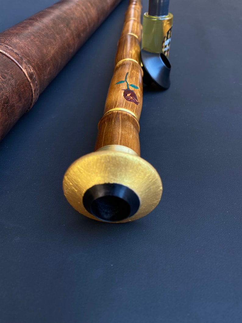 May include: A handcrafted wooden wind instrument with a gold-colored mouthpiece and a rose design. The instrument is accompanied by a brown leather case and a small bottle. The instrument's body is made of bamboo.