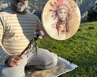 May include: A circular drum with an illustration of a Native American woman wearing a red and brown headdress. The drum is held with a mallet with a purple and black head and a wooden handle. The background features a mountain range and green grass.