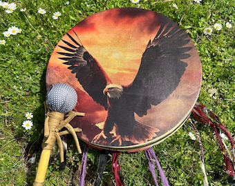 Puede incluir: Un tambor redondo con un diseño de águila, con un fondo de puesta de sol. El tambor va acompañado de una maza con una cabeza de lunares y un mango de madera envuelto en tela color canela. Cintas de colores cuelgan del borde del tambor.