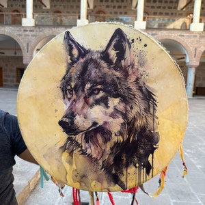 Può includere: Un tamburo rotondo con un ritratto di lupo dipinto. L'opera d'arte presenta una rappresentazione dettagliata della testa di un lupo in tonalità di grigio, nero e marrone. Nastri colorati pendono dal fondo del tamburo.