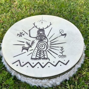 May include: A round frame drum with a cream-colored skin head featuring a black ink illustration of a horned figure, a spiral, a deer, a ram, and a plant. The drum has a thick, white faux fur trim around the circular wooden frame edge.