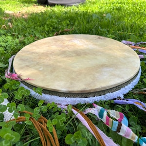 May include: A round, light-colored drum with a decorative border and white fringe, resting on green grass. Colorful ribbons and a drumstick with a white handle and blue accents are also visible. The drum is a musical instrument.