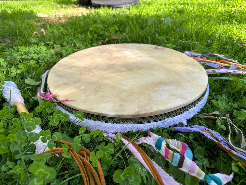May include: A round, light-colored drum with a decorative border and white fringe, resting on green grass. Colorful ribbons and a drumstick with a white handle and blue accents are also visible. The drum is a musical instrument.