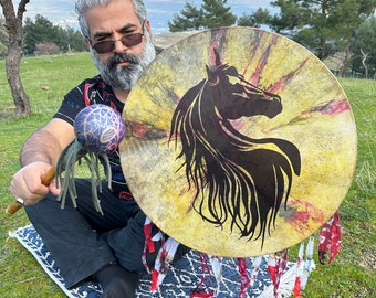 May include: A circular drum featuring a black horse silhouette against a yellow and red backdrop. The drum is adorned with a fringe of red and white fabric. A blue and gold maraca is held in the left hand.