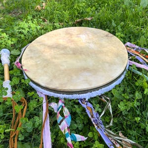 May include: A round, tan drum with a light-colored hide stretched over a wooden frame. The drum is decorated with a white fringe, colorful ribbons, and a wooden drumstick with a white handle. The drum is set on a bed of green grass.