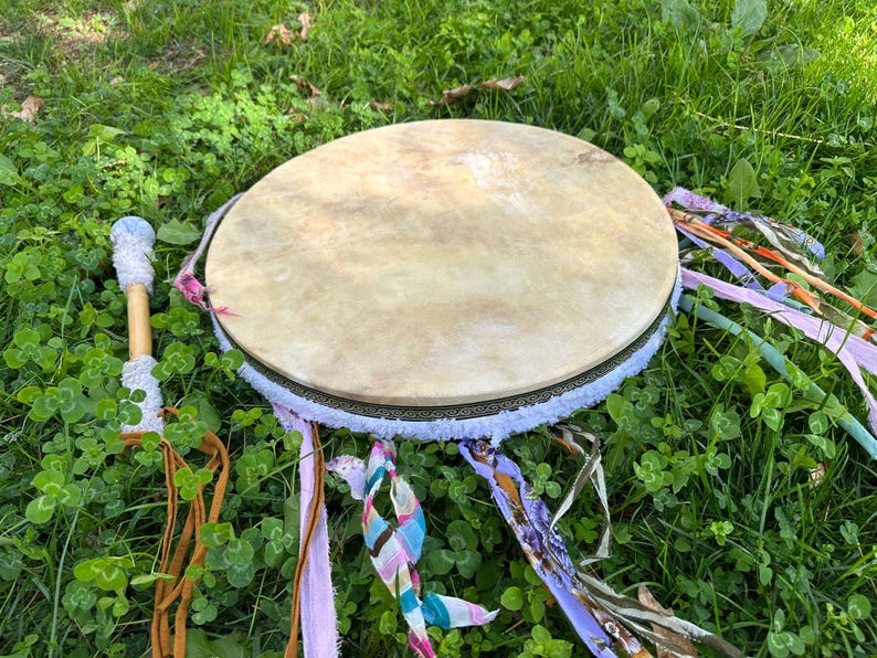 May include: A round, tan drum with a light-colored hide stretched over a wooden frame. The drum is decorated with a white fringe, colorful ribbons, and a wooden drumstick with a white handle. The drum is set on a bed of green grass.