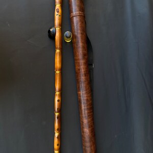 May include: A wooden flute with a black mouthpiece and a rose design, next to a brown leather carrying case. The flute has gold-colored accents and is made of multiple sections. The case has a shoulder strap and a matching cap.