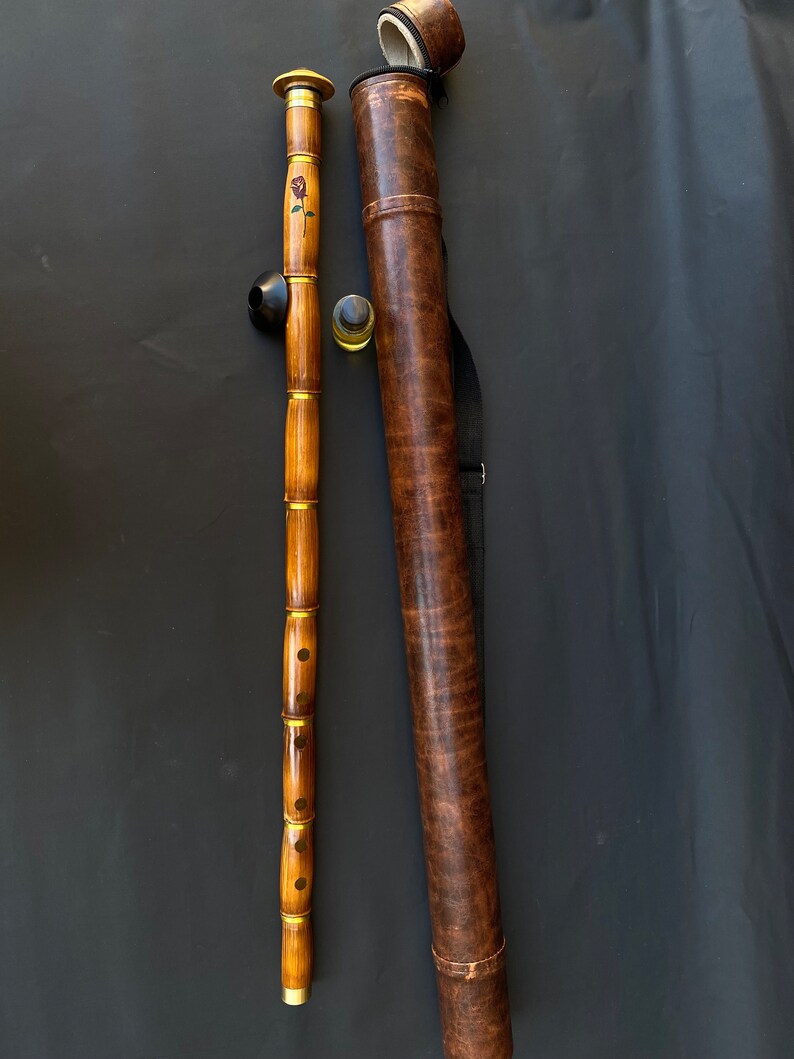 May include: A wooden flute with a black mouthpiece and a rose design, next to a brown leather carrying case. The flute has gold-colored accents and is made of multiple sections. The case has a shoulder strap and a matching cap.
