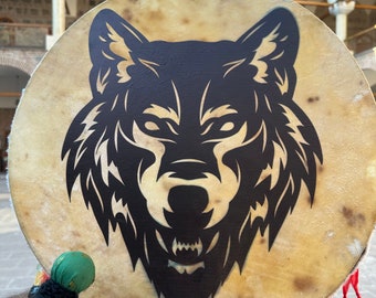 May include: A round, tan drum with a black wolf head design. The drum has a green and black handle and is decorated with red and yellow ribbons. The drum is held up in front of a stone building.