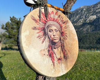 May include: A circular drum featuring a Native American woman's portrait, complete with a feathered headdress, suspended from a tree. The drum is tan with a wooden frame. A drumstick with a black and white head is positioned above.