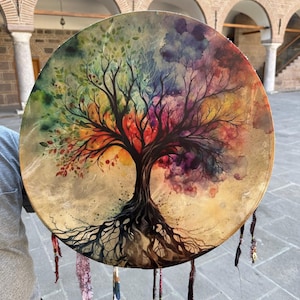 Könnte beinhalten: Eine runde, handbemalte Trommel mit einem lebendigen Lebensbaum-Design. Das Kunstwerk zeigt einen Baum mit bunten Blättern und Wurzeln, vor einem beigen Hintergrund. Dekorative Bänder hängen vom Rand der Trommel.