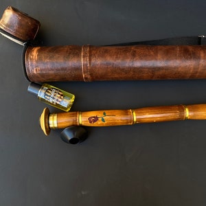 May include: A wooden smoking pipe with a rose design, brass accents, and a black bowl. It is accompanied by a brown leather carrying case and a small bottle with a label. The pipe is approximately 12 inches long.