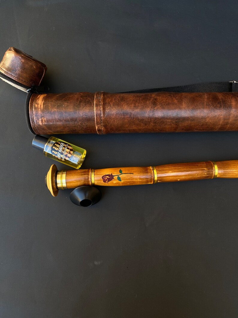 May include: A wooden smoking pipe with a rose design, brass accents, and a black bowl. It is accompanied by a brown leather carrying case and a small bottle with a label. The pipe is approximately 12 inches long.