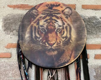 Puede incluir: Tambor redondo con una detallada ilustración de un tigre en tonos marrón, naranja y negro. El tambor está adornado con cintas colgantes de varios colores y texturas, y una pequeña campana. El tambor cuelga contra una pared de ladrillo y piedra.