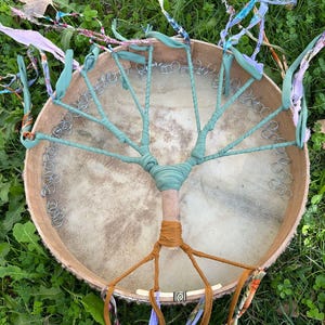 May include: A handmade drum with a tree-like design. The drumhead is tan, with a light brown wooden frame. The tree design is made with teal fabric wrapped around the frame and a central wooden dowel. Colorful ribbons hang from the bottom.