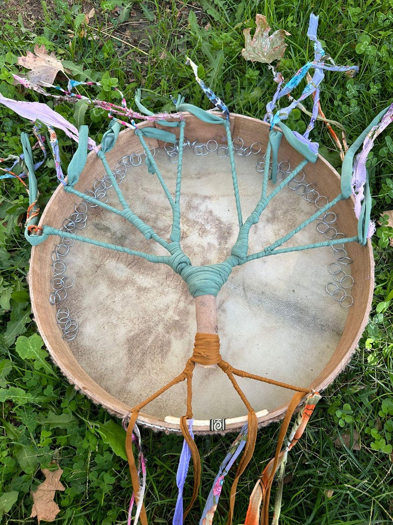 May include: A handmade drum with a tree-like design. The drumhead is tan, with a light brown wooden frame. The tree design is made with teal fabric wrapped around the frame and a central wooden dowel. Colorful ribbons hang from the bottom.