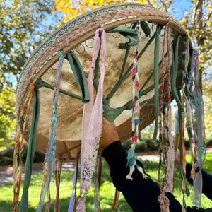May include: A large, round drum with a light brown hide and a decorative wooden frame. The frame is adorned with colorful ribbons and fabric strips in shades of pink, green, and blue. The drum is held up against a backdrop of green grass and trees.
