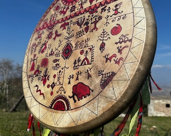 May include: A circular drum with a beige surface, adorned with a complex tribal pattern in red and black. The design includes figures and an elephant. The drum is suspended from a tree, with red and green fabric streamers. A clear blue sky forms the backdrop.