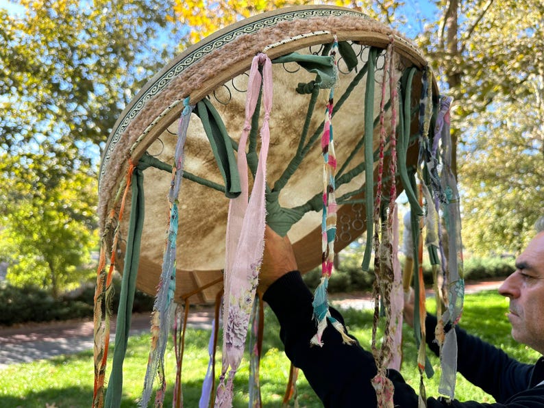 May include: A large, round drum with a light brown hide and a decorative wooden frame. The frame is adorned with colorful ribbons and fabric strips in shades of pink, green, and blue. The drum is held outdoors against a backdrop of trees.