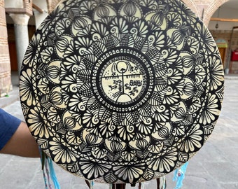 May include: A round, hand-painted drum with a black and white mandala design. The drum features intricate floral and geometric patterns. It has a cream-colored background and is adorned with hanging fabric strips.