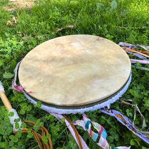 May include: A round, light-colored drum with a natural hide surface, adorned with a decorative border and white fringe. Colorful ribbons and a drumstick with a white, textured head rest on the green grass, suggesting a spiritual or musical theme.
