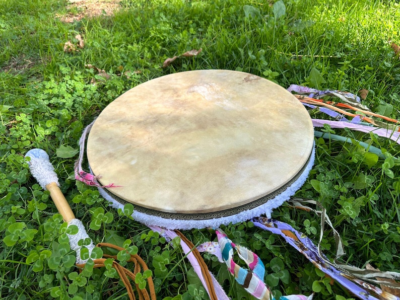 May include: A round, light-colored drum with a natural hide surface, adorned with a decorative border and white fringe. Colorful ribbons and a drumstick with a white, textured head rest on the green grass, suggesting a spiritual or musical theme.