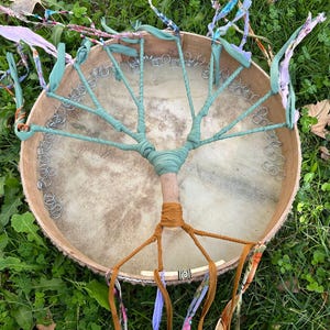 May include: A handmade drum with a tree-like design. The drumhead is a light tan color, with a wooden frame. The tree design is made with teal fabric and brown and orange fabric. A drumstick with a white and blue head is visible.