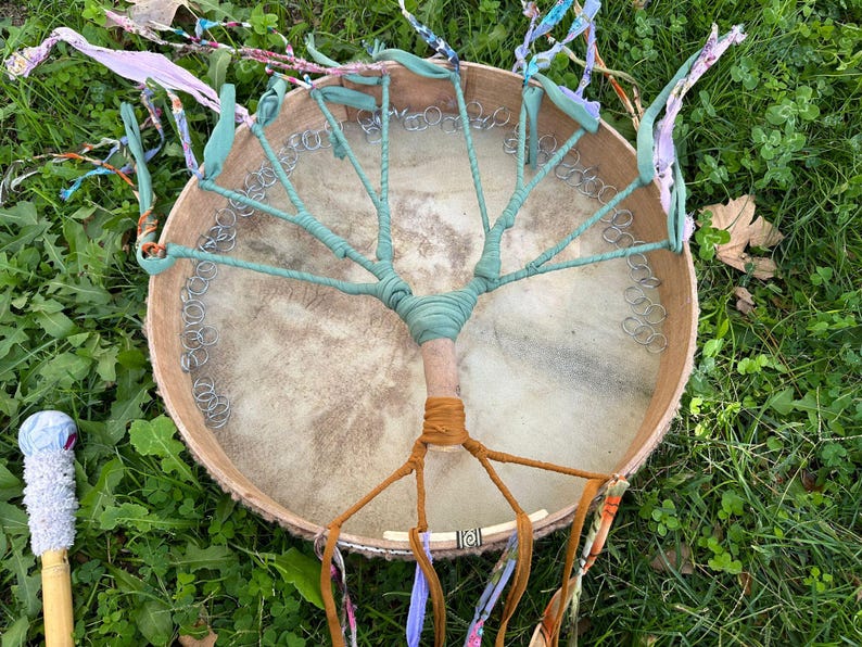 May include: A handmade drum with a tree-like design. The drumhead is a light tan color, with a wooden frame. The tree design is made with teal fabric and brown and orange fabric. A drumstick with a white and blue head is visible.