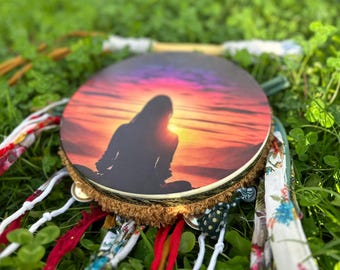 Puede incluir: Un tambor redondo con un diseño de silueta de puesta de sol, rodeado de cintas y campanas de colores. El tambor está sobre un fondo de hierba verde y tréboles. El borde del tambor está adornado con una franja marrón.