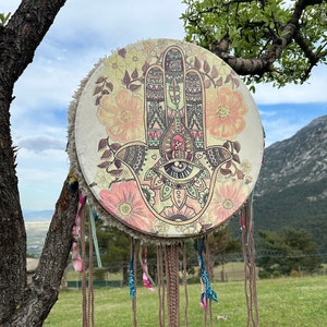 May include: A round, cream-colored drum with a Hamsa hand design, surrounded by floral patterns in pink and peach. The drum is adorned with hanging tassels and ribbons. The background features a tree, green grass, and a mountain range.