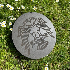 May include: A round, gray stone with a detailed carving of two birds intertwined. The birds have intricate wing and tail designs. The stone is set in a bed of green grass with white daisies.