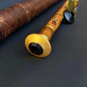 May include: A handcrafted wooden musical instrument with a gold-painted mouthpiece and a rose design. A brown leather case and a small bottle of liquid are also present. The instrument is made of bamboo and has a black mouthpiece.