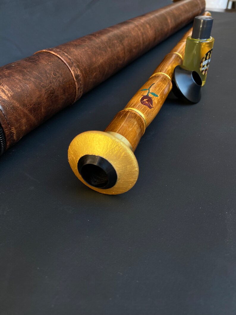 May include: A handcrafted wooden musical instrument with a gold-painted mouthpiece and a rose design. A brown leather case and a small bottle of liquid are also present. The instrument is made of bamboo and has a black mouthpiece.