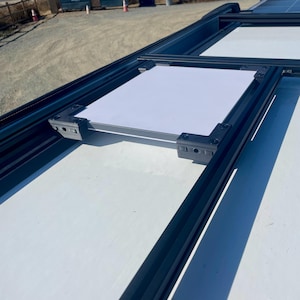 May include: A white solar panel is mounted on a black metal frame on a white surface. The panel is secured with black brackets.