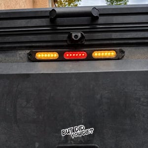 May include: A black vehicle tailgate featuring a horizontal LED light bar with two amber side sections and a red center section. Above the light is a black handle and a camera. A white sticker on the lower section displays the text BUT DID YOU DIE?