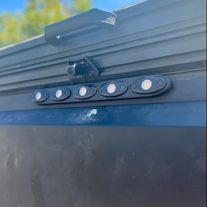 May include: A close-up of a black vehicle component featuring a row of five oval-shaped LED lights with red and white illumination. A small camera is mounted above the lights, and a black handle is visible at the top. The background is a bright blue sky.