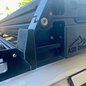 May include: Close-up of a black roof rack with a black metal hinge and a black plastic cover. The cover has the logo "ASG Offroad" embossed on it.