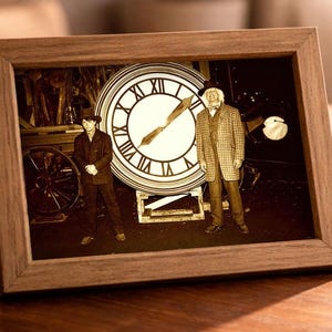 May include: A framed sepia-toned image featuring a clock with Roman numerals and two figures. The frame is made of wood and sits on a wooden table. A lit candle, coffee cup, and books are also on the table.