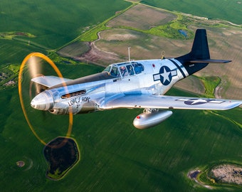 Könnte beinhalten: Ein silbernes und schwarzes P-51 Mustang-Flugzeug im Flug, mit einem sich drehenden Propeller, der eine orangefarbene Unschärfe erzeugt. Das Flugzeug hat einen weißen Stern auf blauem Grund auf den Flügeln und dem Rumpf. Die Worte "LOPEZ' HOPE" sind auf der Nase zu sehen. Der Hintergrund ist ein grünes Feld.