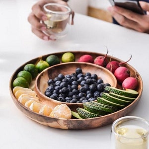 Może przedstawiać: Drewniana taca do serwowania z czterema podzielonymi sekcjami, wypełniona różnymi owocami i warzywami. Taca wykonana jest z naturalnego drewna i ma rustykalny, ręcznie robiony wygląd. Taca idealnie nadaje się do serwowania przekąsek, przystawek lub deserów.