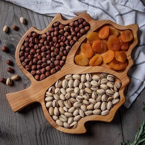 Op de afbeelding: Een houten serveerschaal in de vorm van een blad met drie aparte compartimenten. De compartimenten zijn gevuld met pistachenoten, hazelnoten en gedroogde abrikozen.