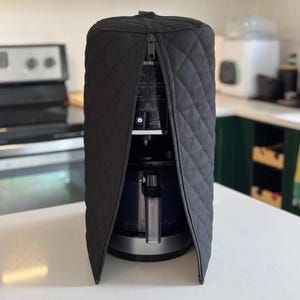 Pode incluir: Uma capa preta acolchoada com um zíper parcialmente aberto, revelando uma cafeteira. A capa é projetada para caber no aparelho, com um padrão de losangos. A cafeteira tem uma base prateada e um reservatório de água transparente.