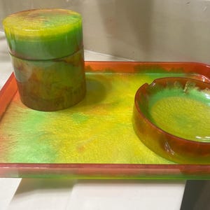 Puede incluir: Una bandeja roja con un diseño de remolino verde y amarillo. La bandeja tiene un cenicero verde y amarillo a juego y un pequeño frasco con tapa.
