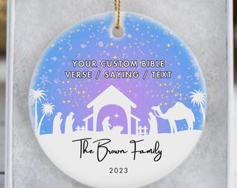 May include: A round Christmas ornament with a Nativity scene in white against a blue and purple gradient background. The ornament has the text "YOUR CUSTOM BIBLE VERSE / SAYING / TEXT", "The Brown Family", and "2023". It is presented in a white gift box.